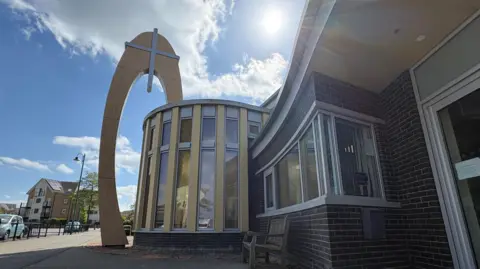 Ben Schofield/BBC A modern, brick-built church, set against a bright sun, in a bright blue and partly cloudy sky. On the left, a crucifix is mounted above a set of tall, vertical windows, built into a bay-type wall, that juts out of the building. On the right is another set of rectangular windows and on the edge of frame is a door leading into the church, though most of that is off-camera. Beyond the church and to the left of the image is a road, low-rise apartment block, tree and street lamp. A couple of cars are parked on the road.
