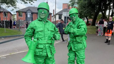Two participants are dressed in bright green military outfits, their glasses and faces are also coloured green and one is holding a loud haler the other a toy machine gun.