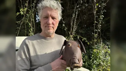 Jo Burn/BBC A man with white, curly hair in a grey jumper. He is holding a leather doll which looks like a dog with a pig's nose.