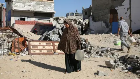 Reuters A displaced Palestinian woman, Makram Hamdouna, carries a water container outside her shelter, in Khan Younis in the southern Gaza Strip December 19, 2024. 