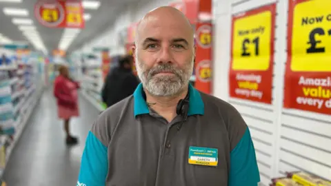 A middle aged man with a balding head and a grey beard looks at the camera as he stands in Poundland. He is wearing a Poundland grey and blue polo shirt and has a name tag with "Gareth - manager" on his top. Two customers can be seen in the aisle behind him.