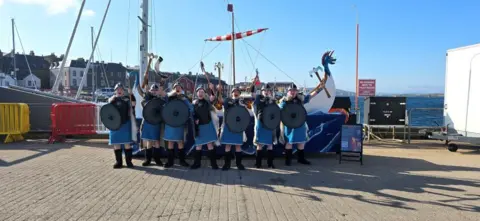 Jacqueline Dawson Seven young boys dressed as Vikings beside a replica long boat. They are dressed in blue and black.