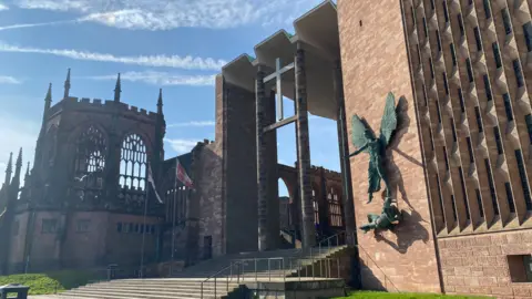 Coventry Cathedral on a sunny day, as seen from University Square. It features the image of St Michael defeating the Devil on the right of the picture and the ruins of the old cathedral on the left