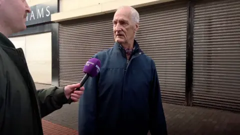 A man in a blue coat is on the high street. A reporter is pointing a microphone at him.