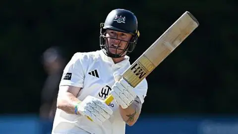 Miles Hammond pulls the ball away through mid-wicket. He is holding his bat high to the right of the picture. He is wearing his whites and has a dak blue helmet on