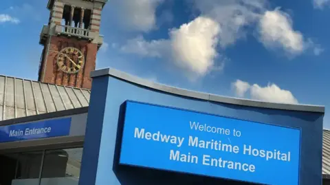 A blue sign reading “Welcome to Medway Maritime Hospital Main Entrance” stands outside the hospital, with part of the main entrance building visible behind it and a red-brick clock tower rising into a blue sky with scattered white clouds.