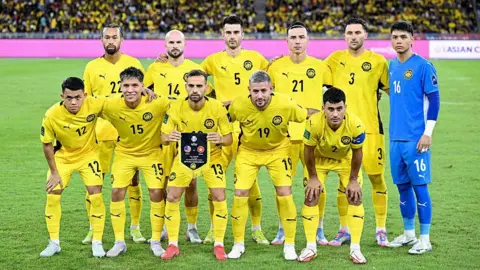 11 football players in yellow jerseys - one goalkeeper wearing blue - pose for a photo on the field