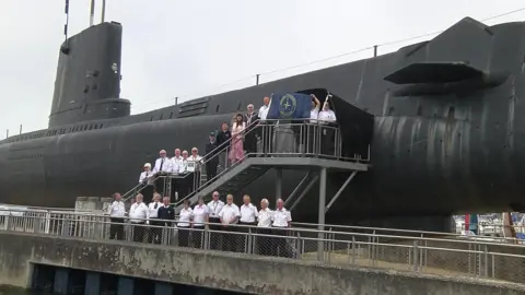 BBC NCI staff outside a submarine holding the blue and gold NCI flag