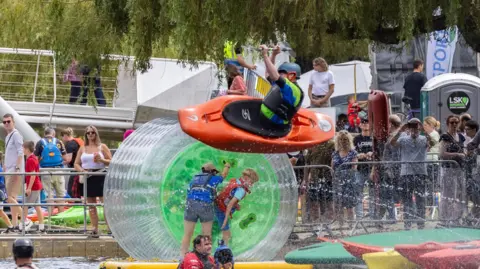 A load of people at a River Festival, taking part in water sports. There is someone in the air in a canoe and people walking over bridges and someone in a zorbing circle.