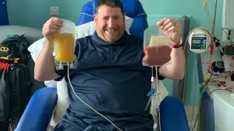 NHS Blood and Transplant Mr Gwilliamin wearing a blue collared t-shirt sitting in a hospital chair holding up a bag of yellow fluid in one hand and a bag containing his donated stem cells in another