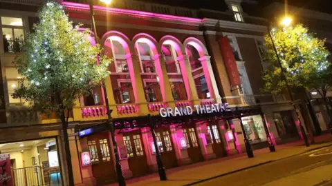 Purple lights shine on the theatre in the dark. The theatre's name is lit up in white and there is a wide entrance with pillars and windows above. Trees are positioned either side of the entrance.