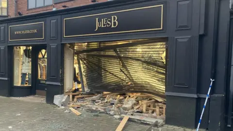 The Jules B fashion clothing shop in Jesmond, Newcastle. The shop front is painted dark grey with gold lettering. The large right-hand window is badly damaged, the window shutters are caved inward and wood and bricks are scattered at the bottom of the window.