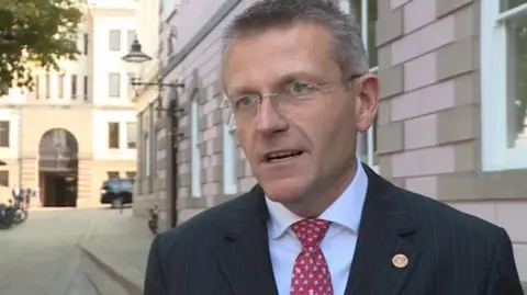 BBC This is a photo of Deputy Ozouf standing outside a building wearing a blue shirt, red tie and a black blazer. He is wearing rimless glasses. 