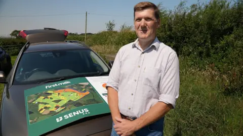 Lee Truelove of First Milk stands in front of a car in a field. On the bonnet is a large cardboard poster showing an example of one of the risk maps the company has produced for each of its farmers. It is a satellite image with fields colour-coded according to risk. Mr Truelove has short brown hair and is wearing a white shirt tucked into light blue jeans