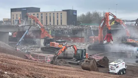 Several orange diggers on a motorway are pictured at work, with a bridge behind them, covered in demolition rubble. Shipping containers are being used to support what is left of the bridge. 