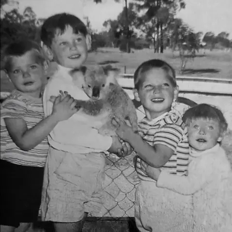 Family Follet, una foto en blanco y negro que muestra a Cheryl Grimmer y sus tres hermanos sosteniendo un koala.