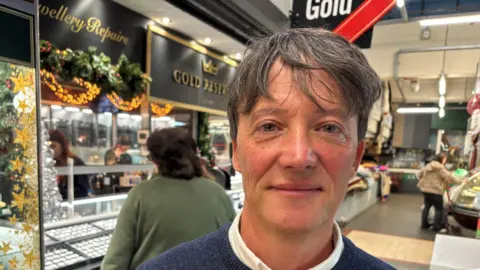 A middle aged man with grey brown hair standing in front of his stall in Swansea market