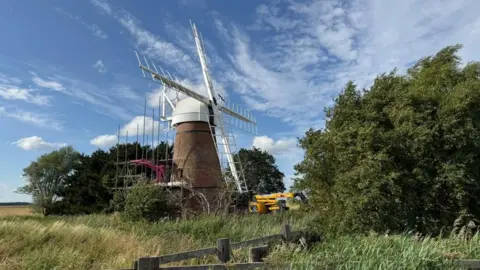 Toft Monks Wind Pump is a brick tower, with a white cap and new sails. Near the tower is a scaffold and a yellow cherry picker. The foreground features reedbeds, with trees on either side, and the sky blue with white cloud.