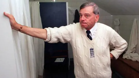 PA Media Bird stands in a back room, with his arm rested on the wall. He is wearing a traditional cricket vest, branding with the ECB logo stitched. He wears a white shirt and tie underneath.