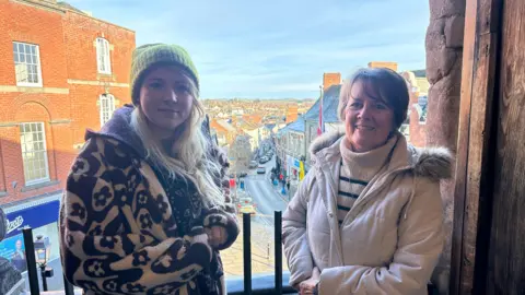Two women stand side by side, smiling at the camera. Behind them is a steep hill lined with shops and houses.