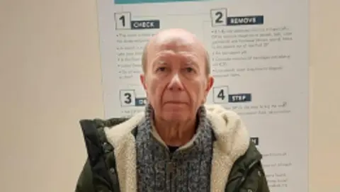 Peter Kindell at a police station: A bald man with grey hair on the side looking ahead. He is wearing a mottled grey jumper with a zip at the neck a green coat which has a white fur lining. He is standing in front of a cream coloured wall. Part of a white poster is visible on the wall. The numbers 1, 2, 3 and 4 are visible on the post plus the words "CHECK", "REMOVE", and "STEP".