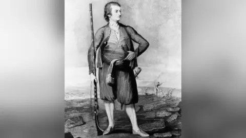 Getty Images A black-and-white historical engraving shows a person standing on a rocky shoreline, dressed in loose traditional clothing and holding a long firearm. The figure is posed upright with one hand on the waist, while a dagger is tucked into a sash around the midsection. In the background, the sea and small boats are visible, suggesting a coastal or maritime setting from an earlier historical period.