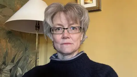 Anna Bailey is looking at the camera. She has a grey bob and is wearing glasses and a dark-coloured winter jumper. In the background is a lampshade, a yellow wall and some patterned wallpaper.