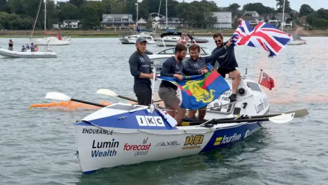 Team United We Conquer - Jack Jarvis, David Bruce, Sam Edwards, and Adam Radcliffe - returning to Hamble from New York. They are on their boat in the water, waving the British flag. Other boats can be seen behind them. It is a cloudy day.