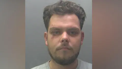 Custody image of Sonny Loveridge, who has brown hair and a short beard. He is wearing a grey sweater and has cuts surrounding his right eye.