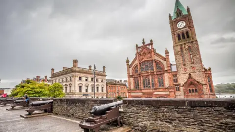 In the foreground on the wall we have several cannons sticking out between the battlements. A girl accompanied by a boy takes pictures of another girl who has climbed a cannon on the wall To the right outside the wall is The Guildhall, building located next to the river, very close to the centre of the city, built in 1887 of sandstone and brick.