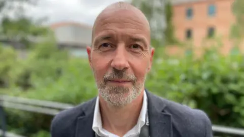 Paul has a beard and is wearing a grey jacket