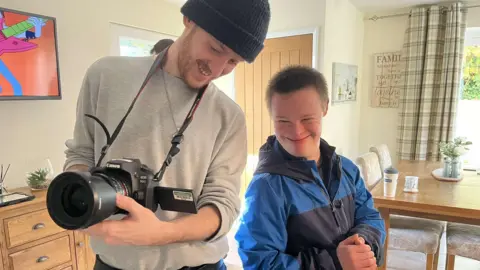 Karina Whelan A man wearing a black beanie hat and a grey jumper is holding a camera and showing another man what is on the screen. They are both looking at the camera and smiling. They are in a kitchen setting.