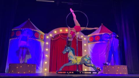 An acrobat, wearing red shorts and top, hanging upside down onto a wheel mounted on a pole, on a stage blue purple and yellow with tents on either side and two other performers standing either side of acrobat.
