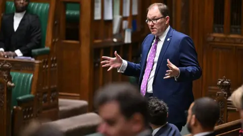 House of Commons Gareth Snell, a man with short hair, glasses, a purple tie and a suit, stands and holds both his hands out while speaking in the House of Commons