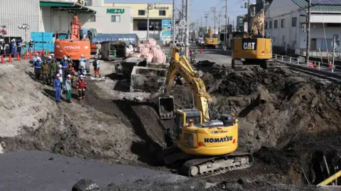 Getty Images An excavator in a sinkhole, as a group of workers with helmets watch on