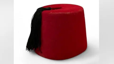 Hanson Auctioneers Fez berada di tengah -tengah gambar di atas meja putih. Ini adalah topi berbentuk bundar merah, setinggi beberapa inci. Ini memiliki rumbai rumbai hitam yang tergantung di sisi kiri memiliki sisi. 