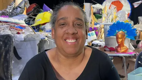 A woman is sat and smiling and behind her are lots of costumes and props for the carnival