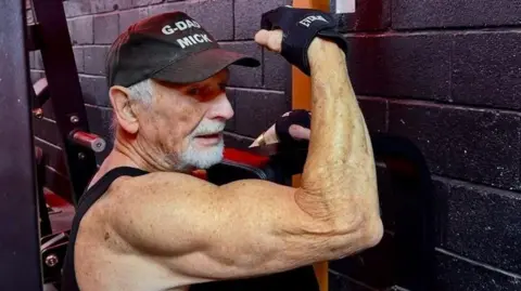 Mick Wadge weighlifting in a gym. His arm is lifted and he has toned biceps.