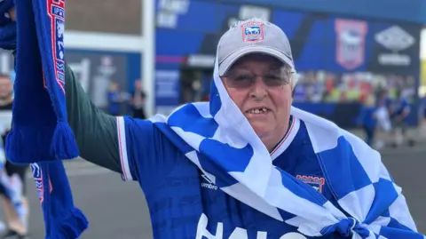 Aimee Dexter/BBC Trevor Loan is standing outside of a football stadium. He has short grey hair and is wearing a blue Ipswich Town football T-shirt, a blue and white flag and is holding a blue scarf.