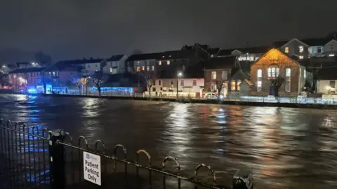 James Wilcox The River Taff in Pontypridd. The water is almost breaking the bank. Police cars and blue lights can be seen in the background on the other side of the river by a row of houses.