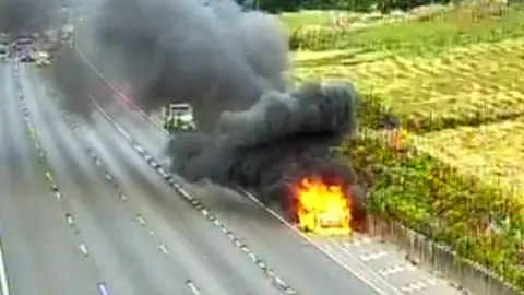 A CCTV image of a vehicle covered in flames on the side of the empty road by a green field. Thick black smoke is rising from the vehicle.