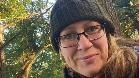 Liz Frost Liz Frost wears rectangular spectacles, a green bobble hat and is smiling. She is in a woods