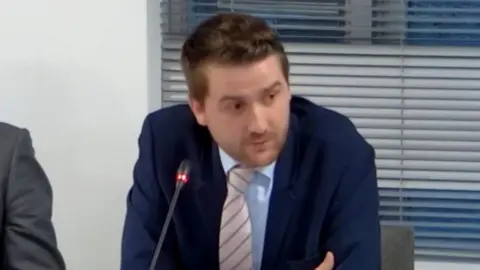 Herefordshire Council Councillor Dan Hurcomb is wearing a blue suit and shirt with a light-coloured tie and speaking into a microphone.