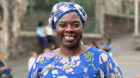 Penny Marie is smiling at the camera wearing a blue dress and a head wrap.