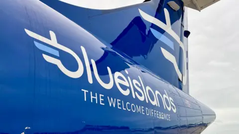 The tail of a plane, in royal blue. It says 'Blue Islands,The Welcome Difference' in white writing. Above the writing, there is a white and light blue logo. To the right of the image, the sky is grey.