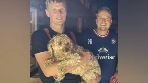 A firefighter holding a dog with a man smiling next to him.