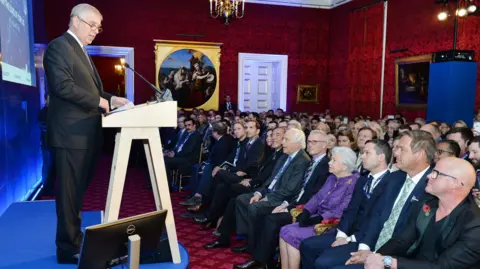 PA Media Prince Andrew stands at a lectern in a grey suit, speaking to an audience including the Queen, who wears a purple outfit, David Stern, and other celebrities and influential people including Heston Blumenthal