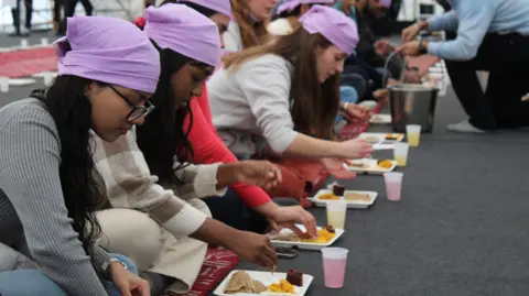 Young women sitting in a line with lilac head scarves on. They are sitting cross legged on the floor and are reaching towards white plates with vegetarian food on them. They have plastic cups near them filled with juice