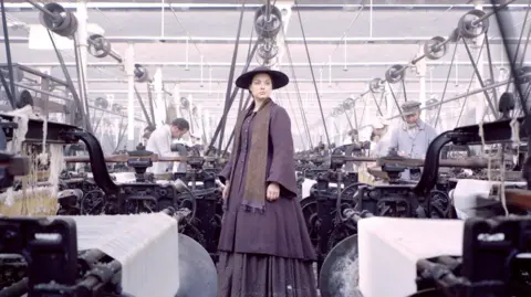 Actress Daniela Denby-Ashe as Margaret Hale in the BBC drama North and South. She is standing in a purple hat, long coat and dress between the mill's looms with extras performing as mill workers weaving white cloth. 