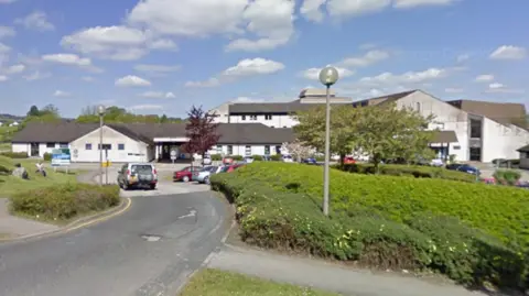 Google Westmorland General Hospital in Kendal. White buildings, situated in the countryside, are surrounded by hedgerows.